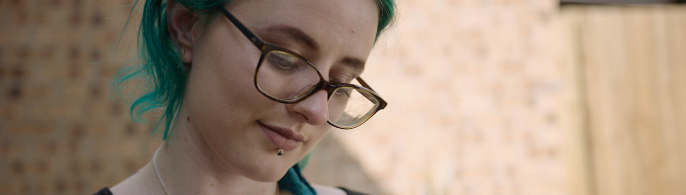 Person with green hair and glasses, sitting in front of a wall and reading a book.