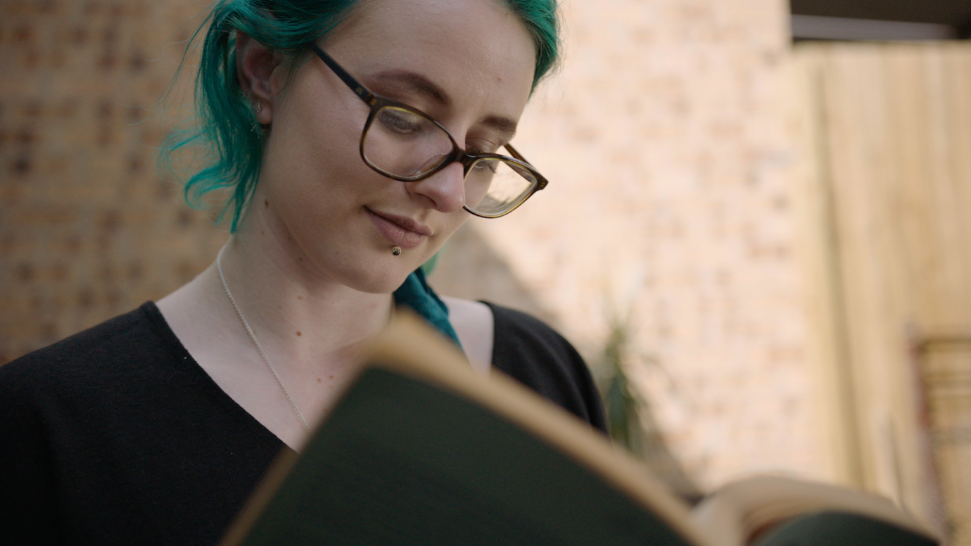 Person with green hair and glasses, sitting in front of a wall and reading a book.