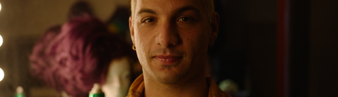 Man with short bleached hair and a check shirt sitting in front of a dressing table mirror.