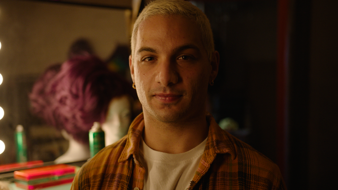 Man with short bleached hair and a check shirt sitting in front of a dressing table mirror.