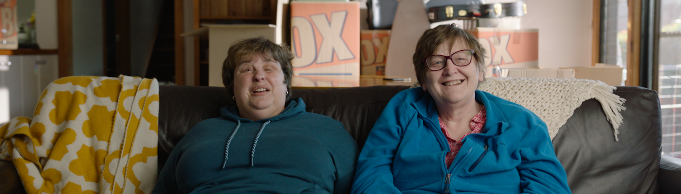 Two women wearing hoodies and trousers sitting on a sofa with moving boxes in the background.