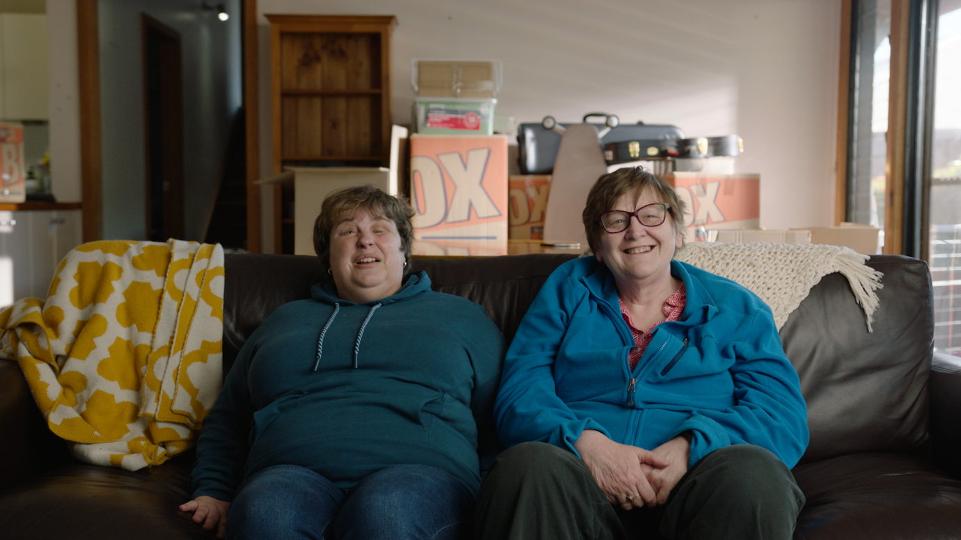 Two women wearing hoodies and trousers sitting on a sofa with moving boxes in the background.