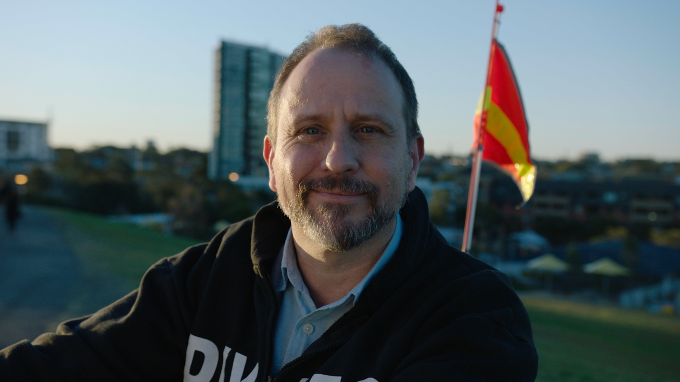 Man with short hair and goatee sitting on the crest of a hill. There is a safety flag and buildings in the background