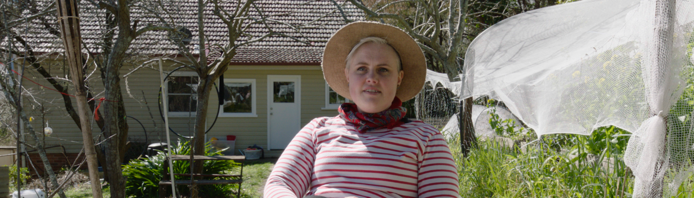 Woman in a straw hat and striped top sitting on a chair in a garden with trees and a single story house in the background.