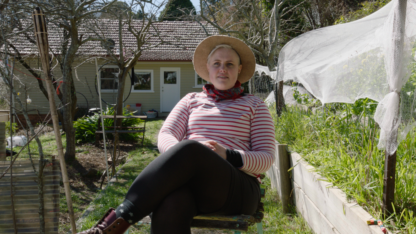 Woman in a straw hat and striped top sitting on a chair in a garden with trees and a single story house in the background.