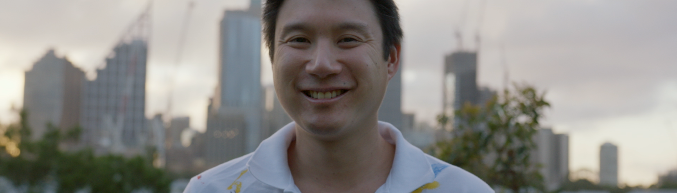 Man in a white polo shirt standing outside with trees in the background and the Sydney skyline in the distance. 