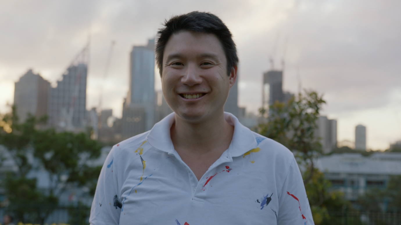 Man in a white polo shirt standing outside with trees in the background and the Sydney skyline in the distance. 