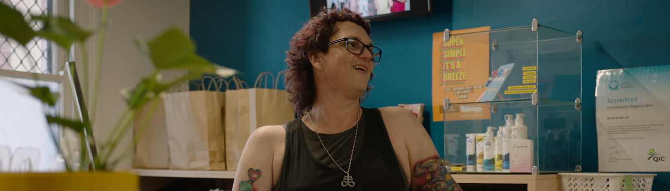 Woman wearing a black sleeveless top, glasses and pendant stands smiling at a reception desk.