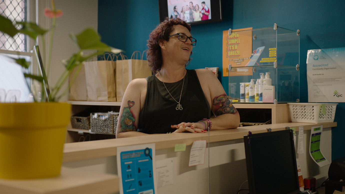 Woman wearing a black sleeveless top, glasses and pendant stands smiling at a reception desk.